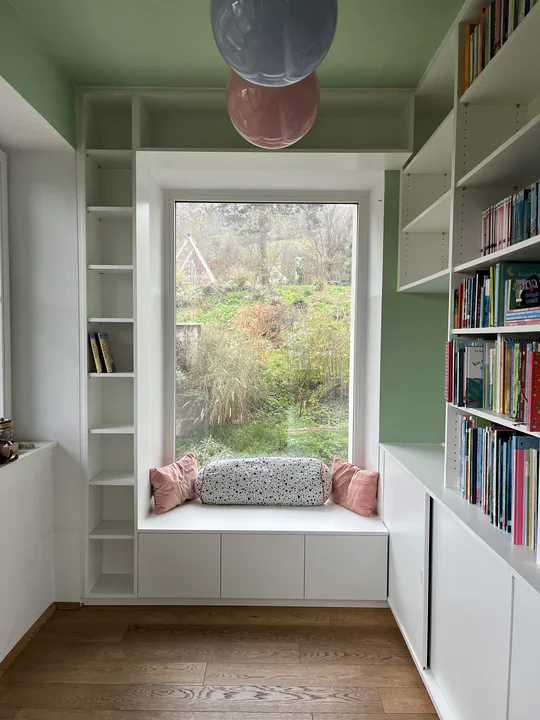 Bibliothekszimmer mit gefüllten Regalen und einer gepolsterten Sitzfläche auf der Fensterbank