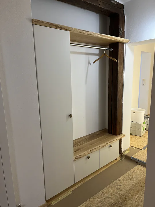 Weißer Garderobenschrank neben einer offenen Nische mit Kleiderstange, Hutablage und Bank aus Holz mit Baumkante