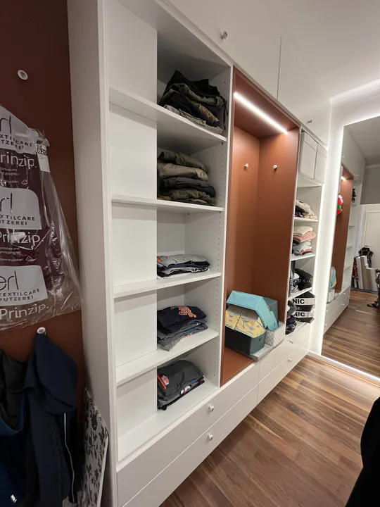 Garderobe mit offenen weißen Fächern, beleuchteter Nische und rostroter Rückwand über dunklem Holzboden