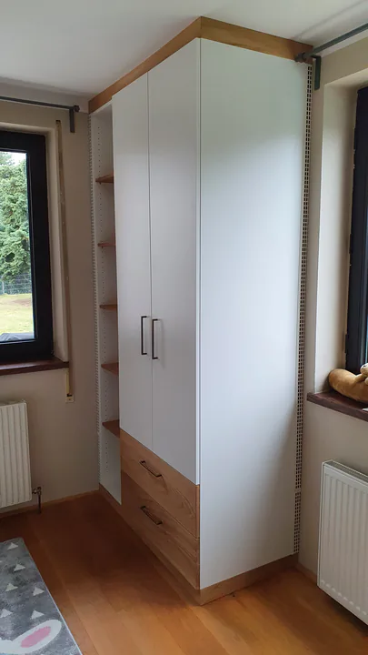 Weißer Hochschrank zwischen zwei Fenstern, mit Laden aus Holz im Unterbau und offenen Fächern an der Seite
