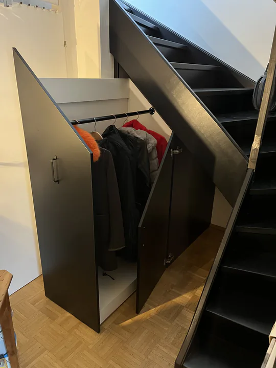 Schwarzer Einbauschrank unter einer Treppe, Türen offen, innen hängt Garderobe