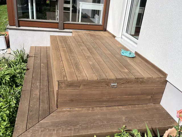 Dieselbe Holzterrasse von der Seite, die breite Stufe führt in einen dicht bepflanzten Garten
